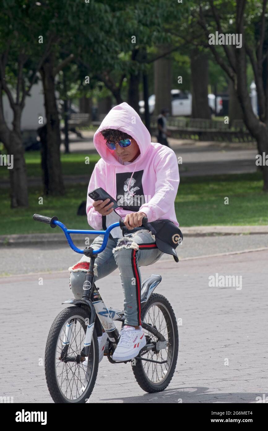 Un jeune homme à capuchon rose lit depuis son téléphone tout en pédalant sa bicyclette. À Flushing Meadows Corona Park, Queens, New York. Banque D'Images