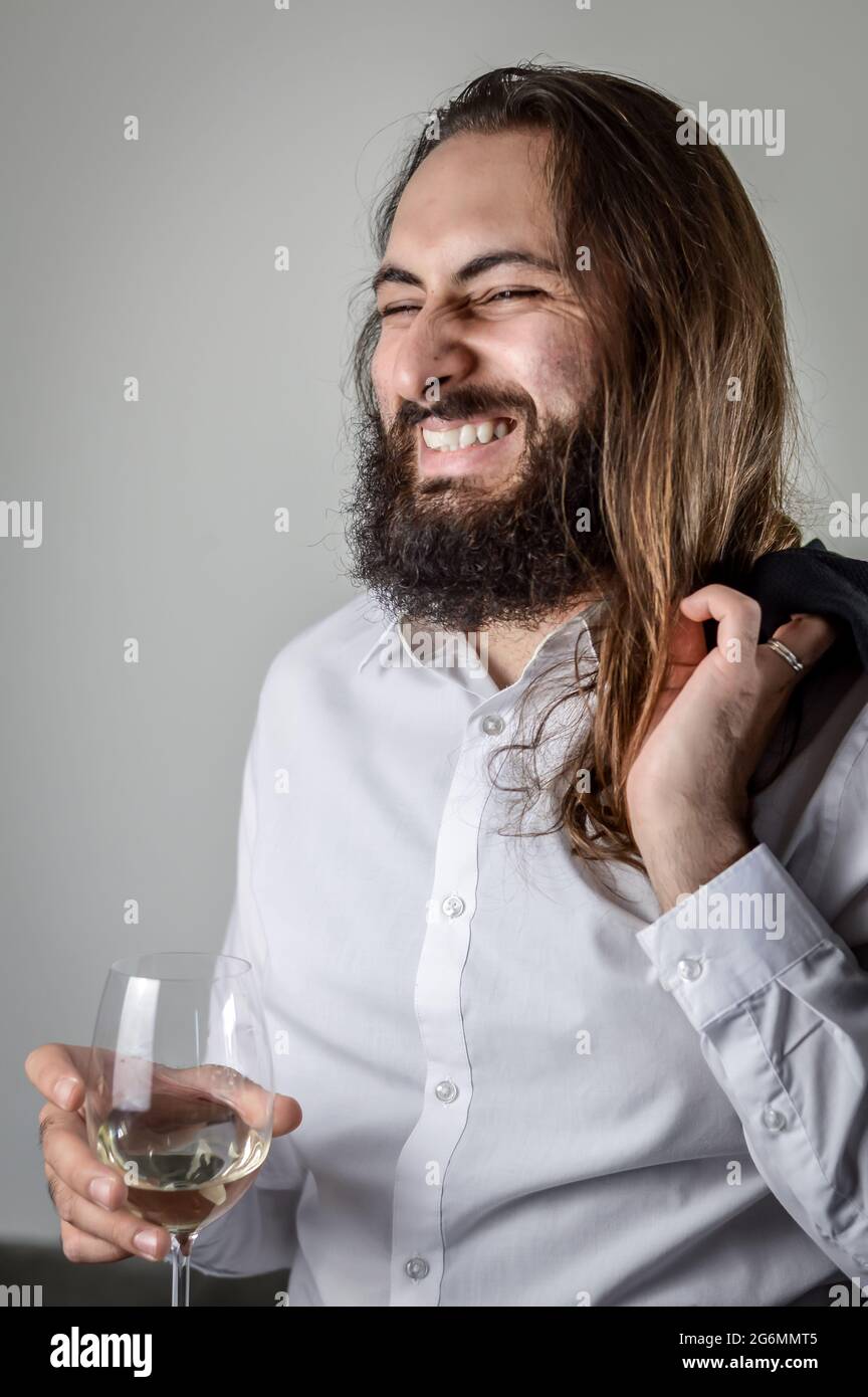 Portrait d'un jeune homme d'affaires du Moyen-Orient avec une barbe et de longs cheveux riant gaiement tout en tenant un verre de vin Banque D'Images