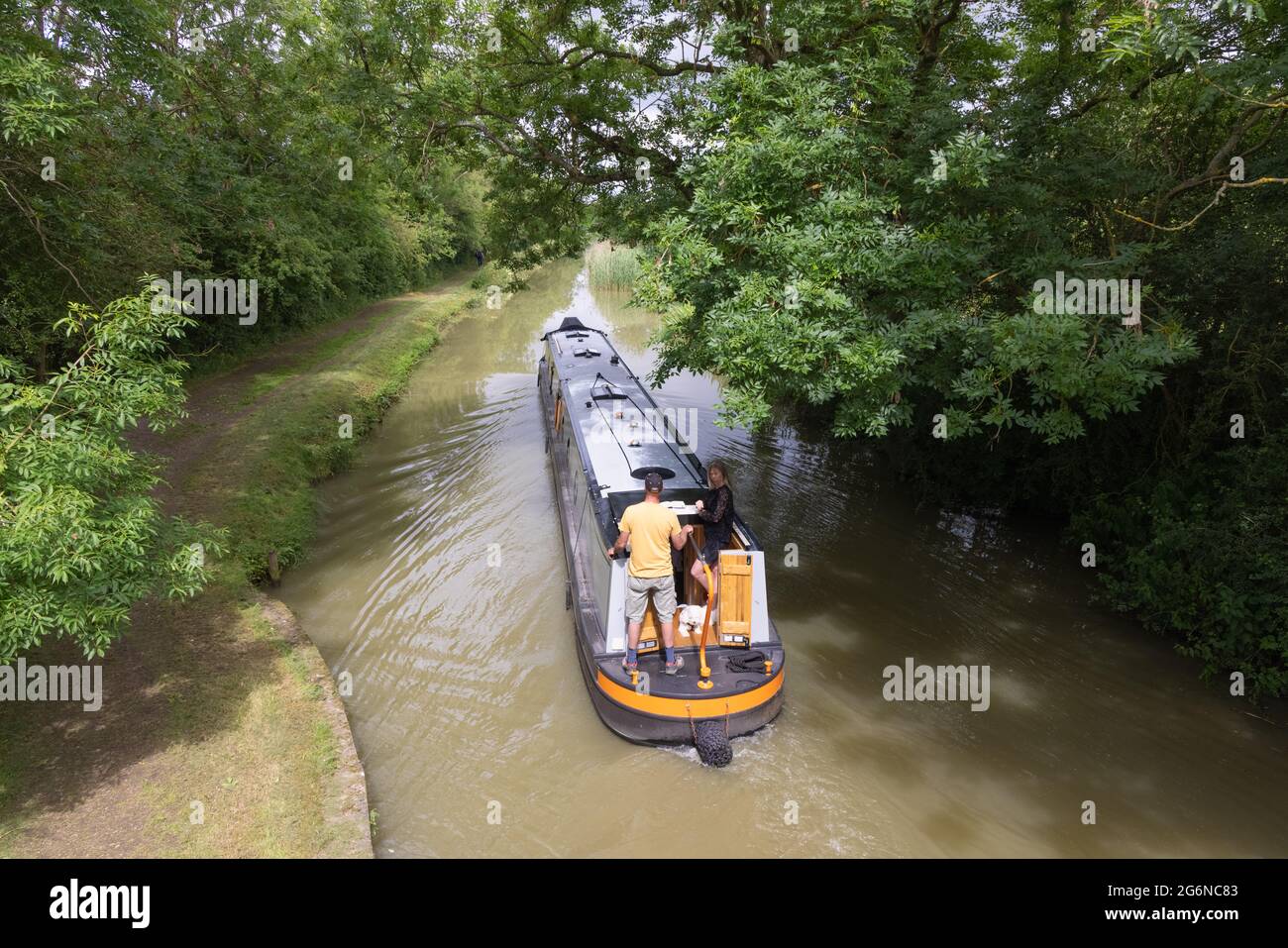 Crick, Northamptonshire, Royaume-Uni, 7 juillet 2021 : un couple avec un petit chien à la poupe d'un bateau à rames navigue sous les arbres le long du canal de Grand Union. Banque D'Images