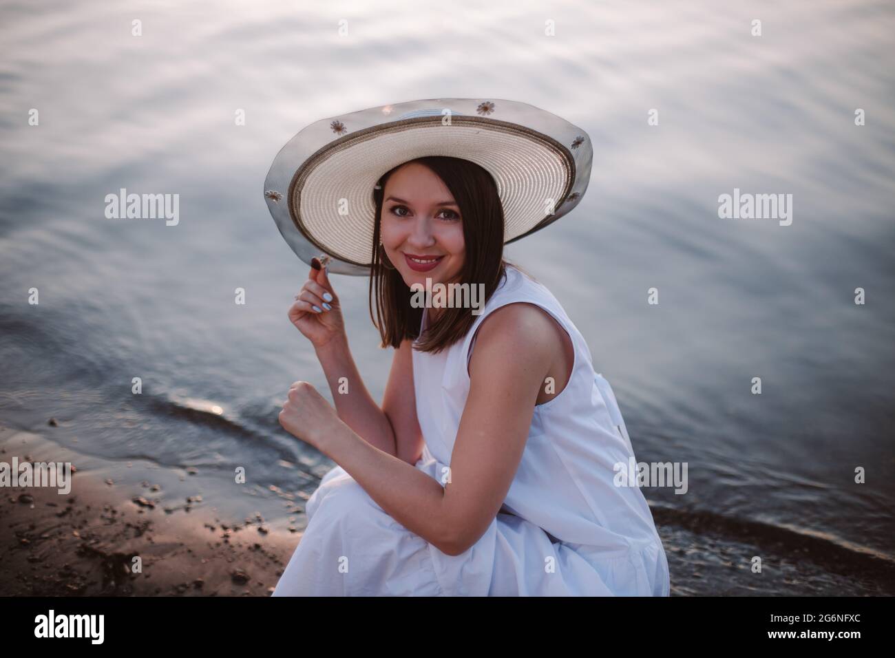 un portrait romantique d'une fille rieuse dans une sunress blanche et un chapeau de paille sur une date sur la plage au bord de la rivière ou de la mer au coucher du soleil Banque D'Images