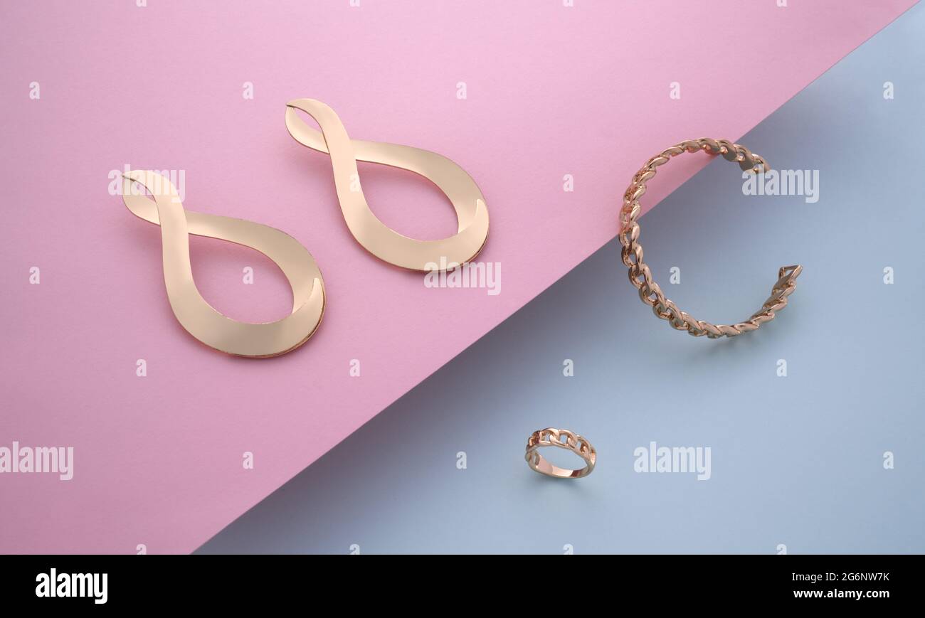 Bracelet et bague en forme de chaîne avec paire de boucles d'oreilles dorées sur fond rose et bleu Banque D'Images