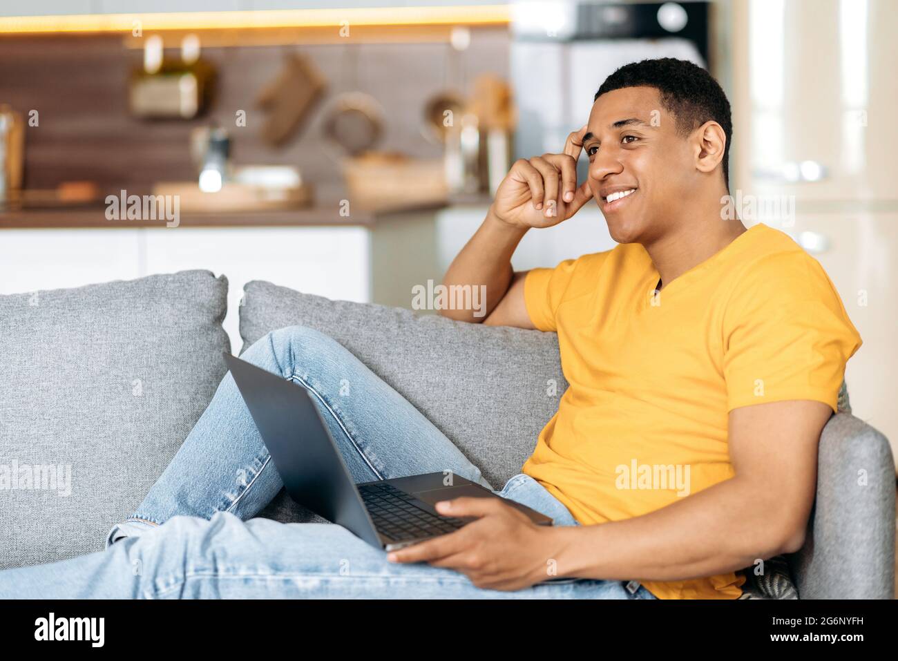 Positif latino gars assis sur un canapé à la maison dans le salon, à l'aide d'un ordinateur portable, la navigation sur Internet, chat en ligne sur les réseaux sociaux, fait des commandes en ligne, regardant à côté, souriant Banque D'Images