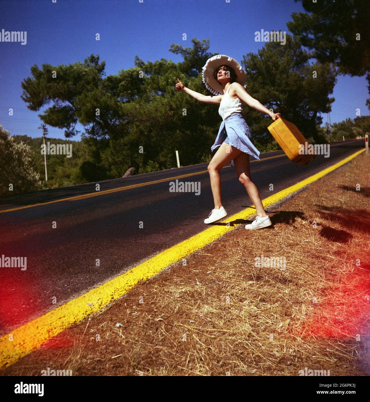 20s âge une femme avec un chapeau de paille, Jean short, t-shirt en tricot et valise jaune hitchranking au bord de la route du pays. Prise de vue avec un objectif à double réflexe A. Banque D'Images