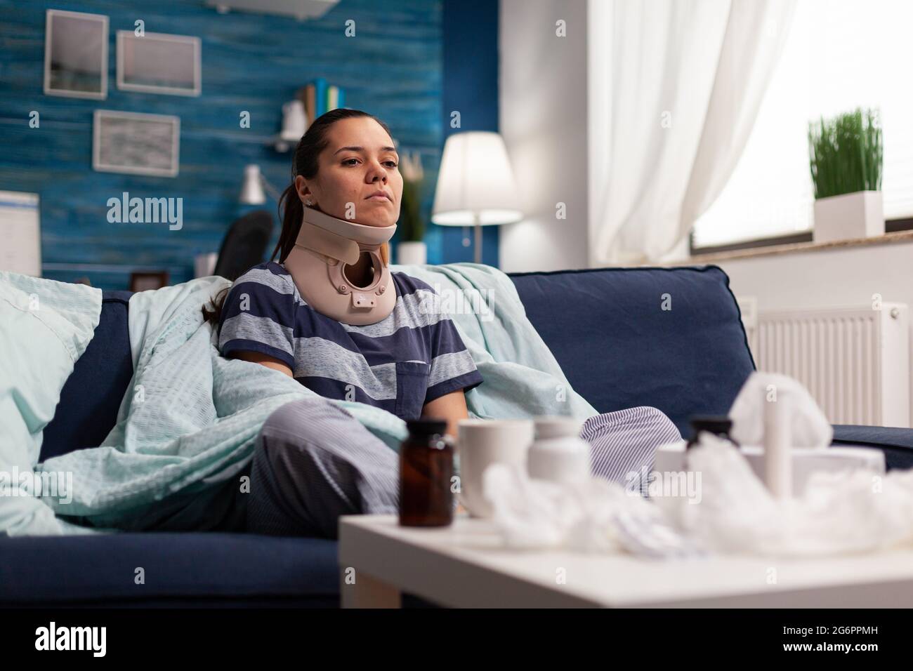 Femme avec col cervical assis sur un canapé souffrant de douleur de colonne vertébrale. Jeune caucasien avec une blessure physique prenant un médicament contre la douleur après un accident du dos, avec une gêne Banque D'Images