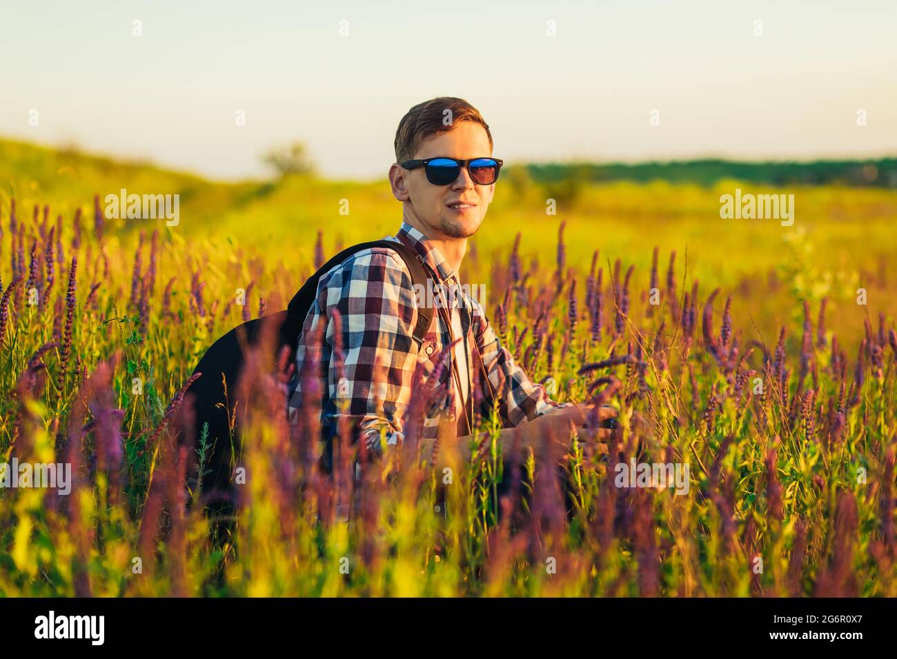 Jeune homme en lunettes de soleil assis dans un champ avec des fleurs sauvages, homme à la campagne pendant le coucher du soleil, appréciant la nature, concept de voyage, nature Banque D'Images