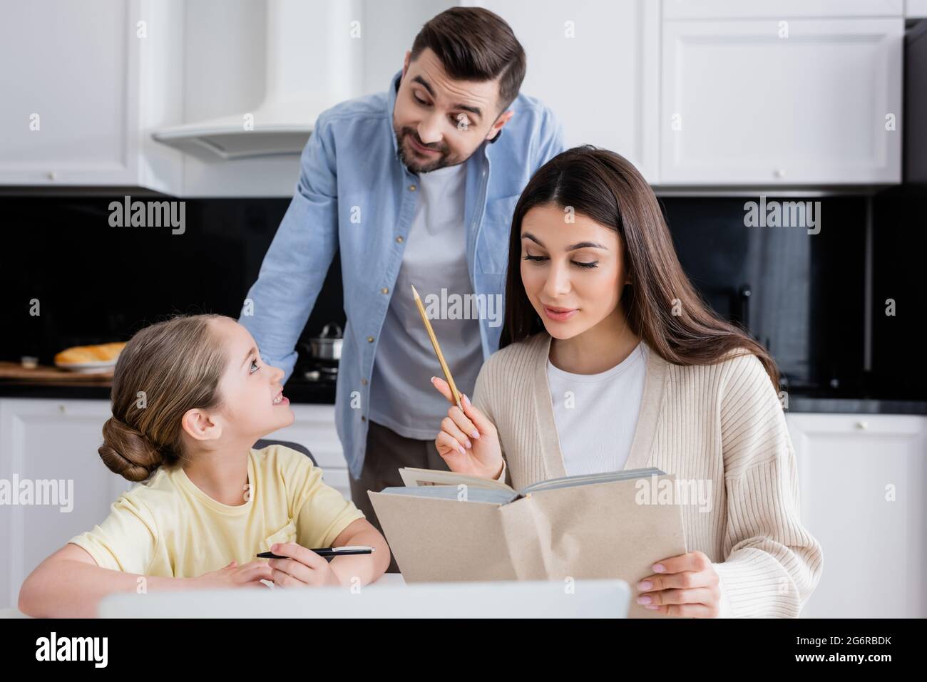 homme souriant regardant la fille faire ses devoirs avec maman dans la cuisine Banque D'Images