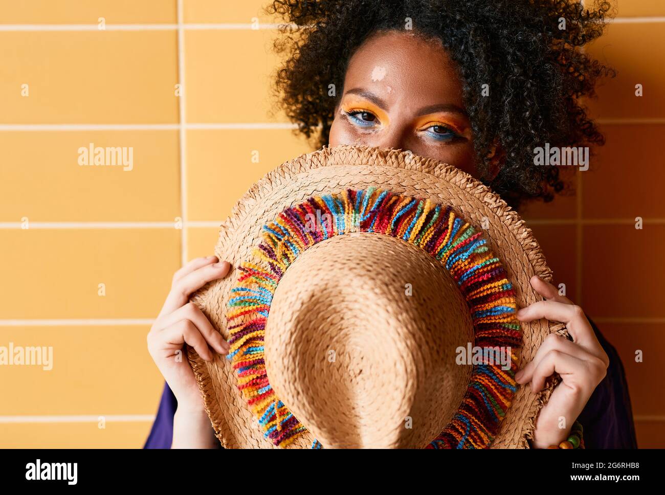 Femme afro-américaine avec chapeau de paille coloré devant le visage ayant un maquillage brillant et élégant debout près du mur orange et regardant l'appareil photo. Bonjour Banque D'Images