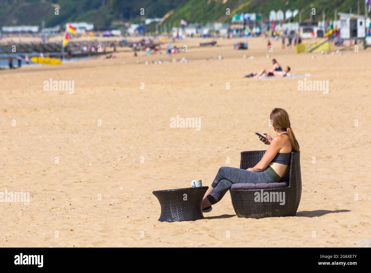 Bournemouth, Dorset, Royaume-Uni. 9 juillet 2021. Météo au Royaume-Uni : belle journée ensoleillée, mais humide sur les plages de Bournemouth, tandis que les amateurs de soleil se dirigent vers le bord de mer pour profiter du soleil. Femme assise sur une chaise en osier avec une tasse sur une table en osier sur le sable, tenant un téléphone portable smartphone. Crédit : Carolyn Jenkins/Alay Live News Banque D'Images