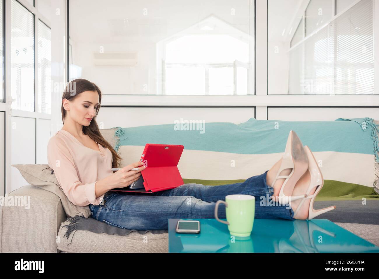 Longueur pleine vue latérale d'une jeune femme créative assise sur un canapé avec ses pieds sur une table à café, tout en travaillant sur le comprimé dans le bureau d'un c Banque D'Images