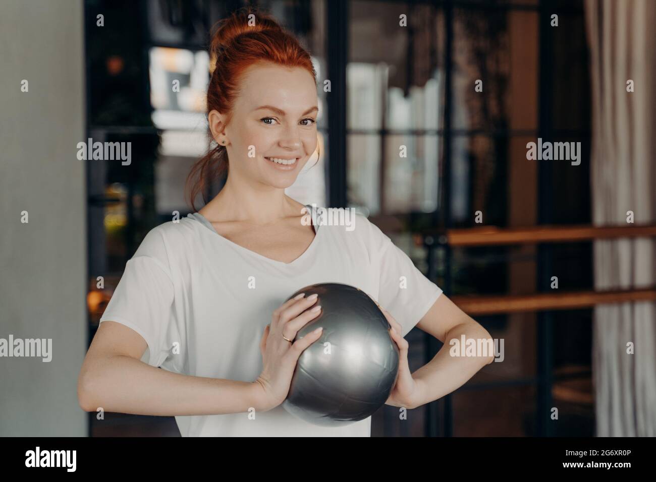 Belle femme de gingembre dans les vêtements de sport souriant à l'appareil photo tout en tenant argent petite taille de fitball dans les mains Banque D'Images