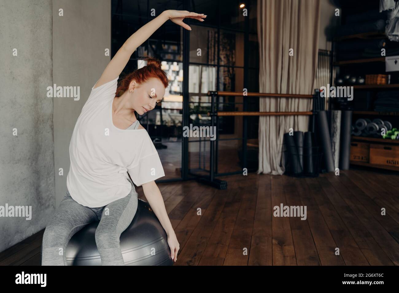 Femme rouge sportif aux cheveux assise sur un grand fitball et un corps qui s'étire avant de s'entraîner dans la salle de gym Banque D'Images