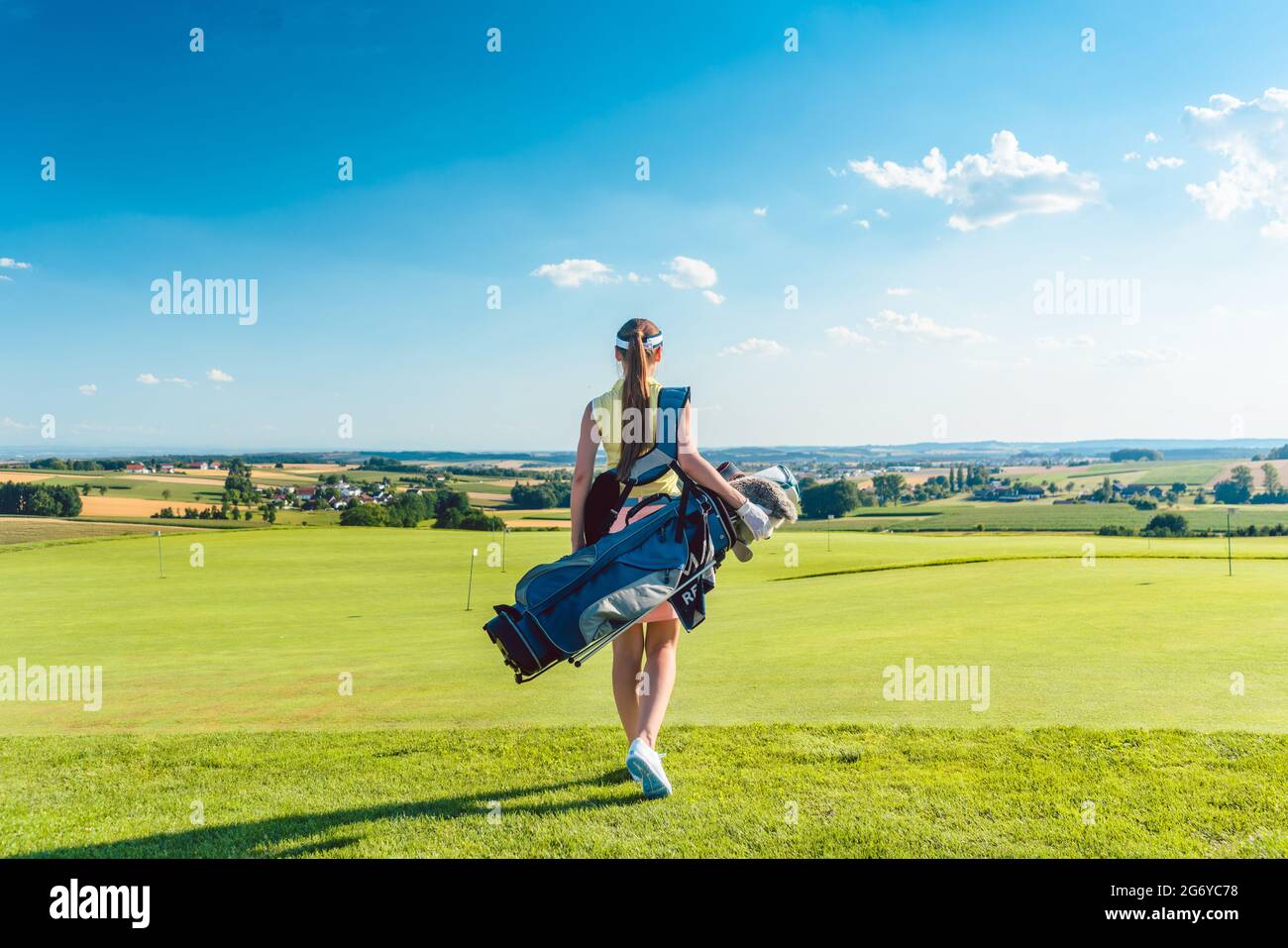 Vue arrière sur toute la longueur d'une femme active portant un sac de stand bleu, tout en marchant sur l'herbe vers une aire d'entraînement pour le golf professionnel en un soleil Banque D'Images