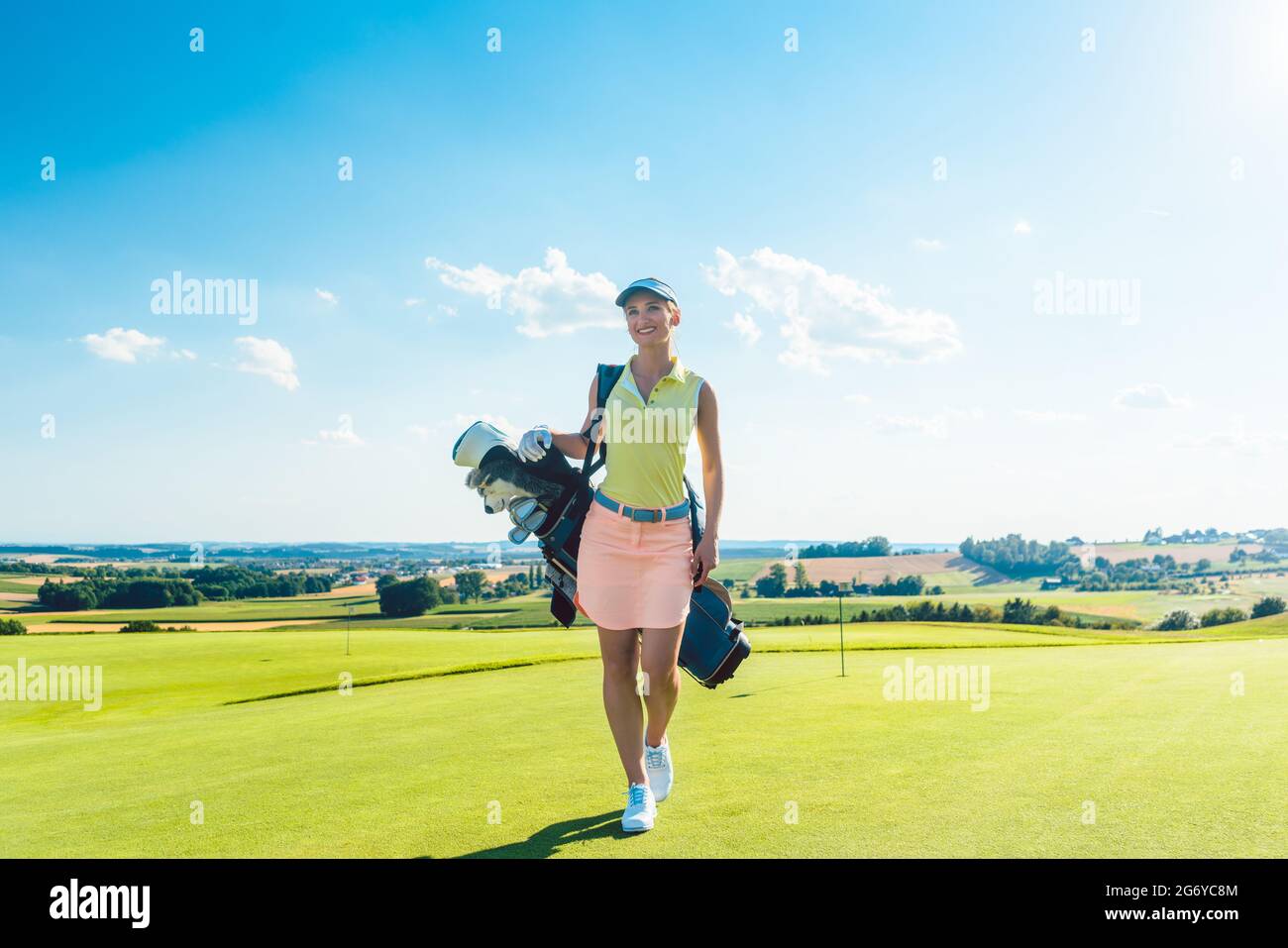 Vue arrière sur toute la longueur d'une femme active portant un sac de stand bleu tout en marchant sur l'herbe, vers une aire d'entraînement pour le golf professionnel en un soleil Banque D'Images