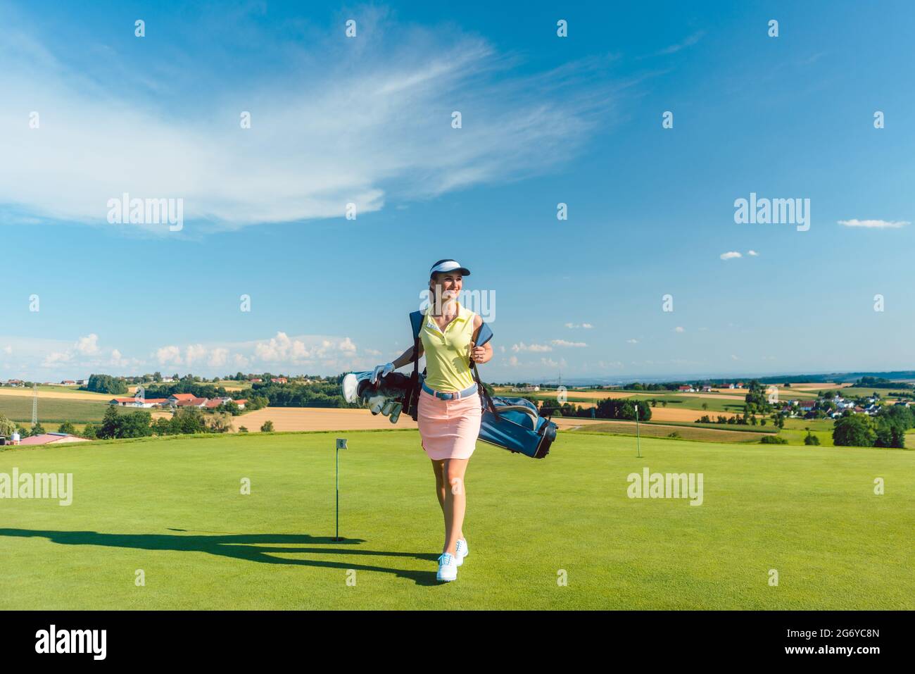 Vue arrière sur toute la longueur d'une femme active portant un sac de stand bleu, tout en marchant sur l'herbe vers une aire d'entraînement pour le golf professionnel en un soleil Banque D'Images