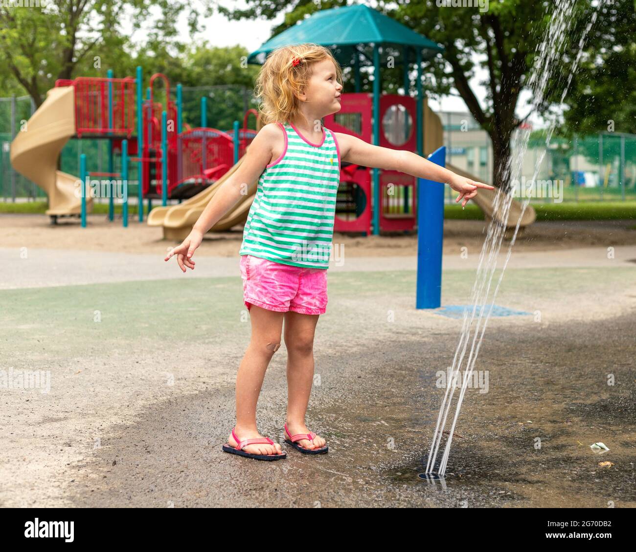 Un petit enfant souriant joue avec de l'eau dans le parc public local le jour d'été chaud. Petite belle fille s'amuser à la fontaine Playgrou Banque D'Images