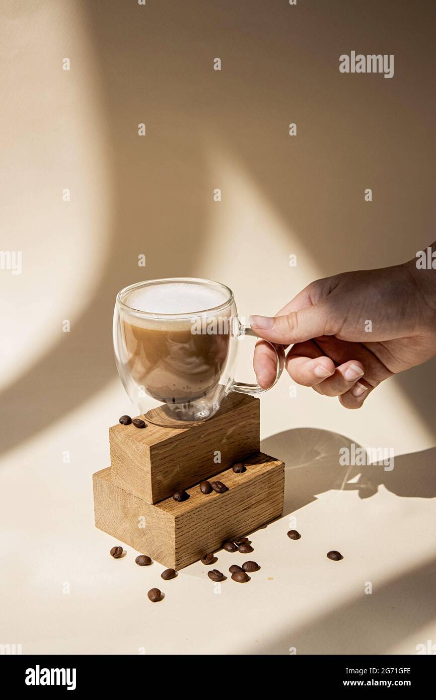 Café dans une tasse de verre sur fond beige. La main tient une tasse. Des photographies stylées et minimalistes à la lumière du jour avec ombres et lumière. Banque D'Images