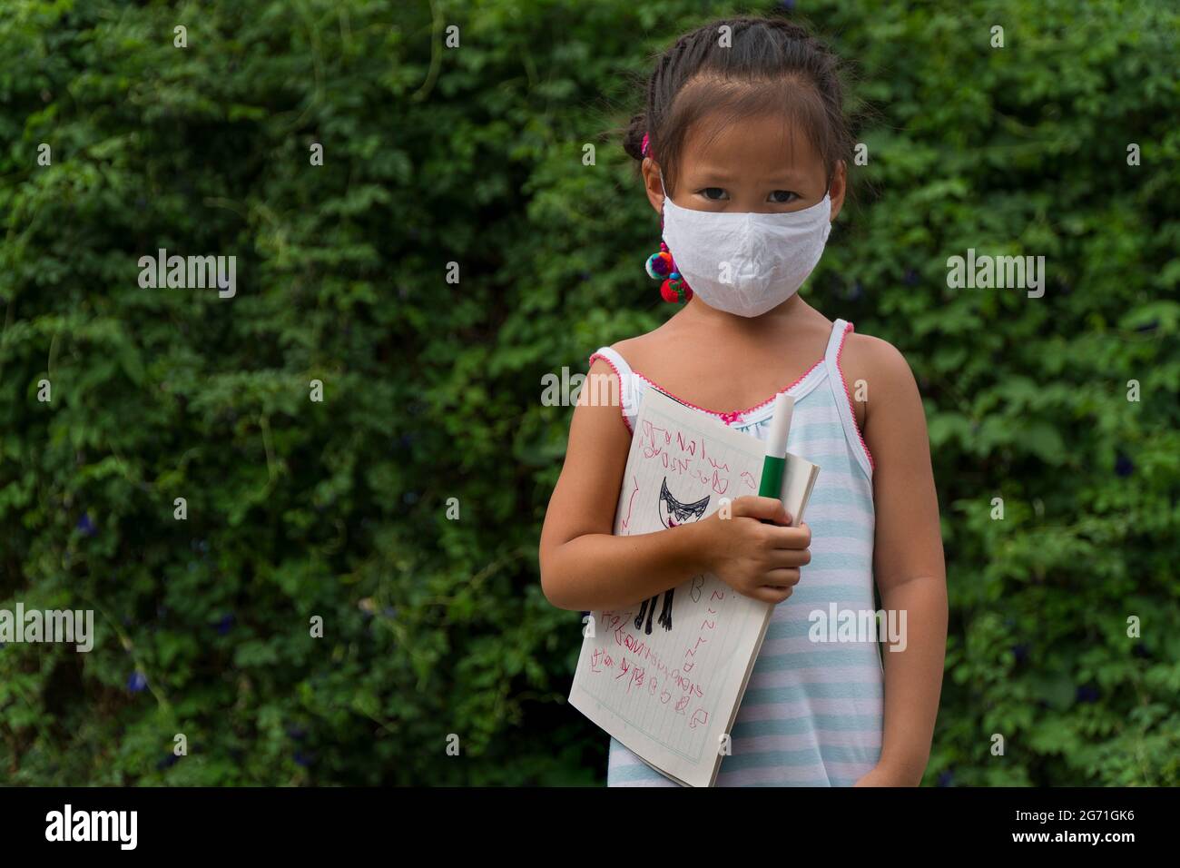 Petite fille asiatique portant un masque facial avec sac à dos vert tenant la papeterie dans le fond vert du parc naturel. Concept de retour à l'école. Banque D'Images
