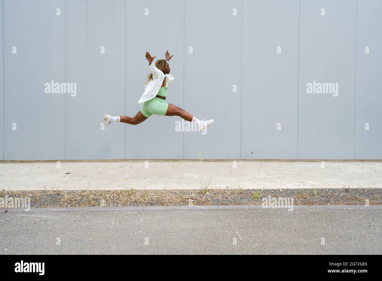 Portrait complet d'une femme afro-américaine sautant avec les jambes écarées et les bras étendus. Femme en vêtements de sport et blouson blanc avec braid jaune Banque D'Images