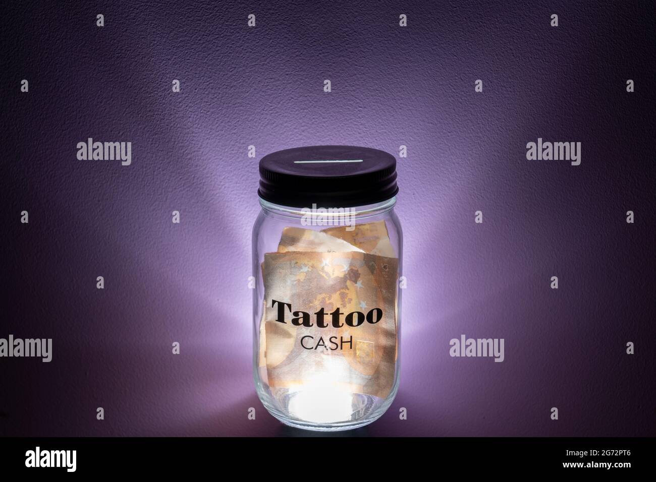 Pot en verre avec inscription « Tattoo CASH » pleine de billets en papier sur fond éclairé Banque D'Images