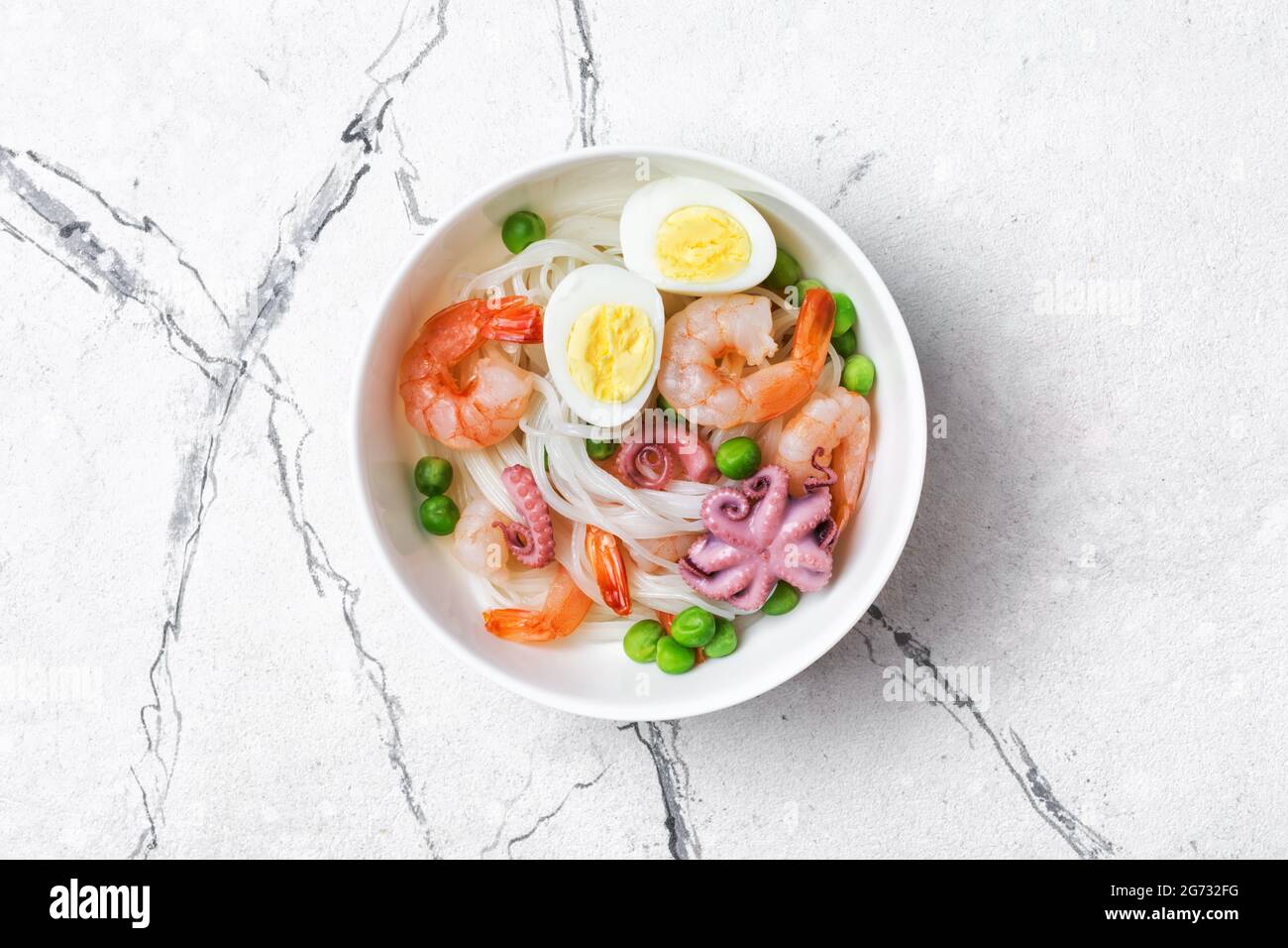 Vue de dessus d'un bol avec des nouilles de riz chinoises, de savoureuses crevettes, du bbe octopus, des œufs de poulet et des pois verts sur fond de marbre blanc Banque D'Images