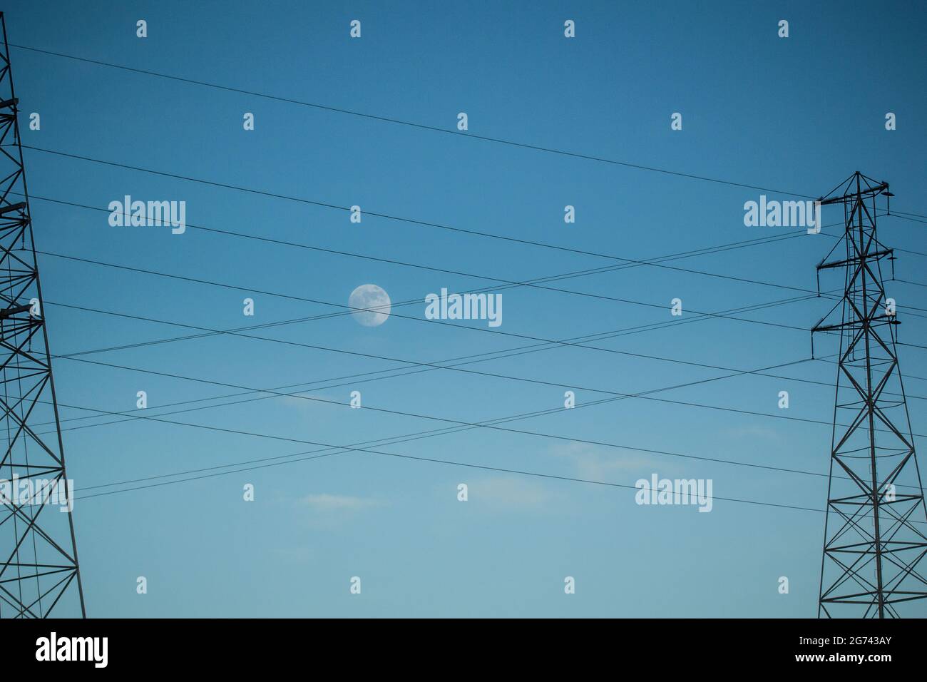 La lune par temps clair derrière une croix de câbles d'alimentation suspendue à deux tours de transmission en réseau en Californie. Banque D'Images