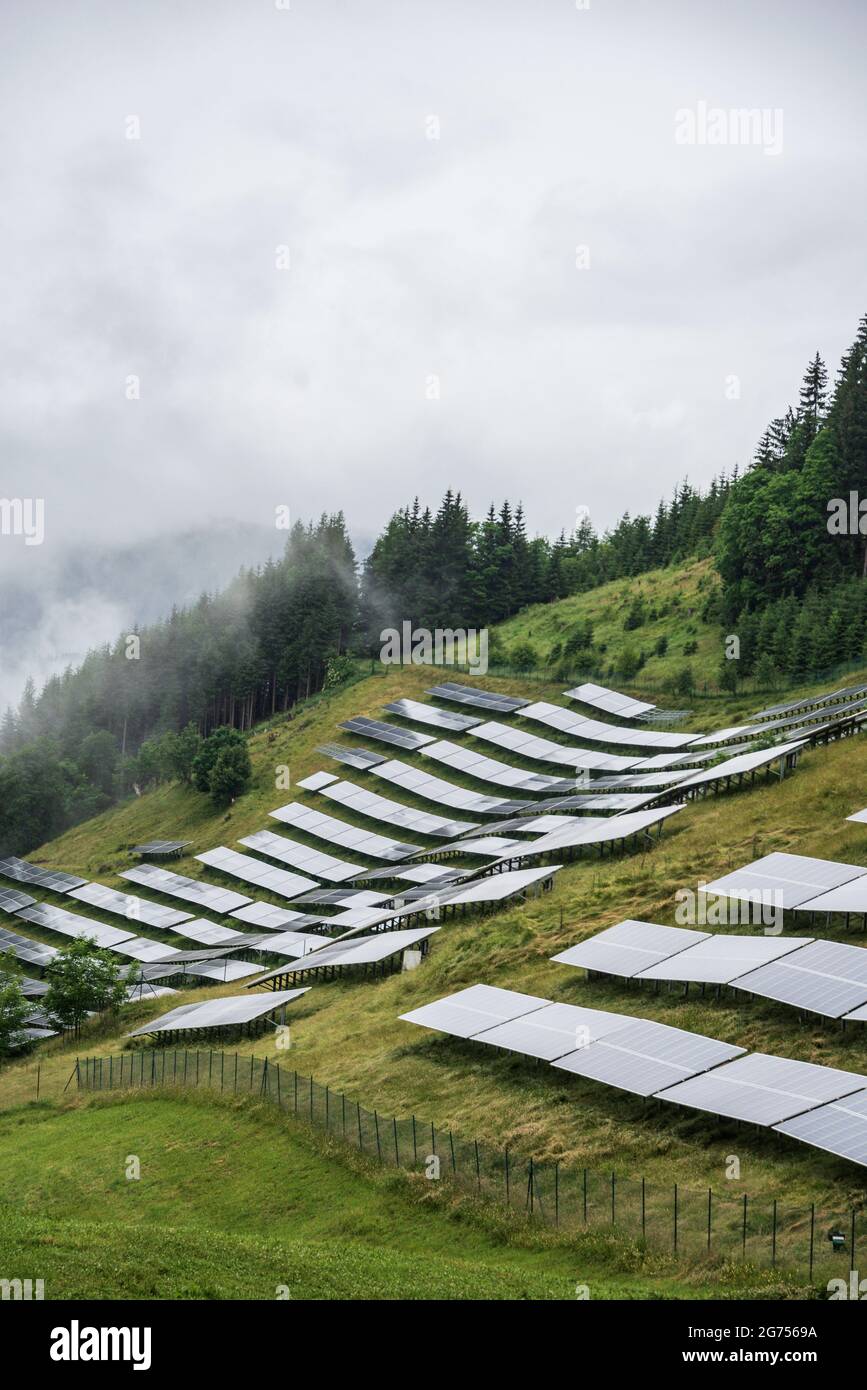 Centrale solaire photovoltaïque située sur une pente de montagne dans les Alpes. Jour brumeux, herbe verte de pâturage et ciel bleu et les montagnes créent un frais Banque D'Images