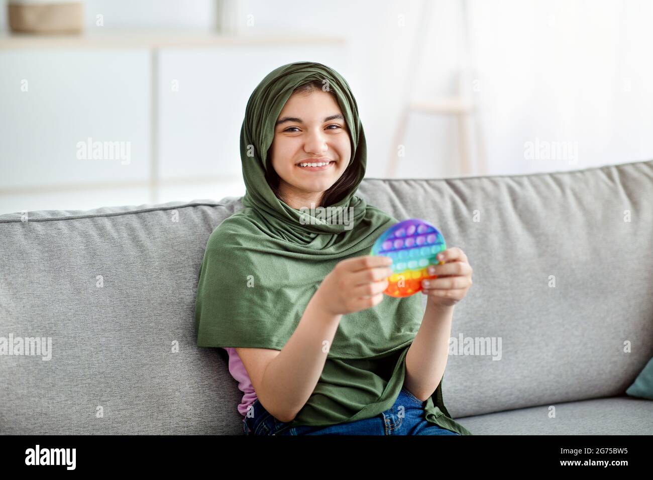 Jeune fille indienne souriante dans hijab assis sur un canapé confortable, jouant avec le jouet pop IT tendance à la maison Banque D'Images