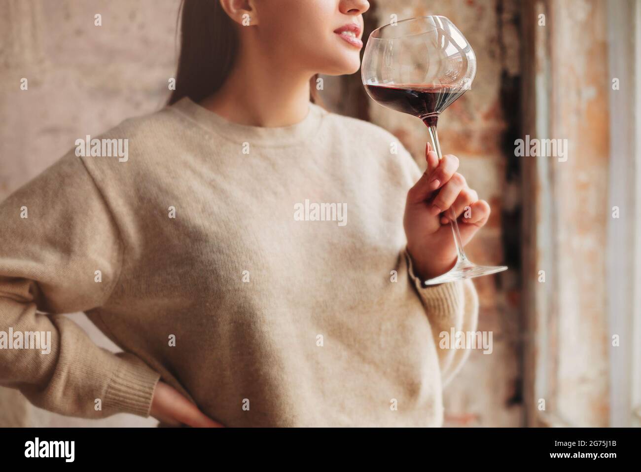 Crop Happy female en chandail sentant le vin rouge dans un grand verre sur fond gris Banque D'Images