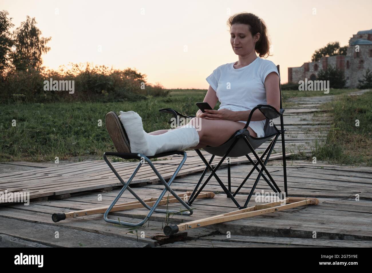 Femme avec cassé dans le casting est assis sur une chaise dans la nature et est chantant par téléphone. Blessures aux jambes. Perte de mobilité. Réadaptation après une fracture. Banque D'Images