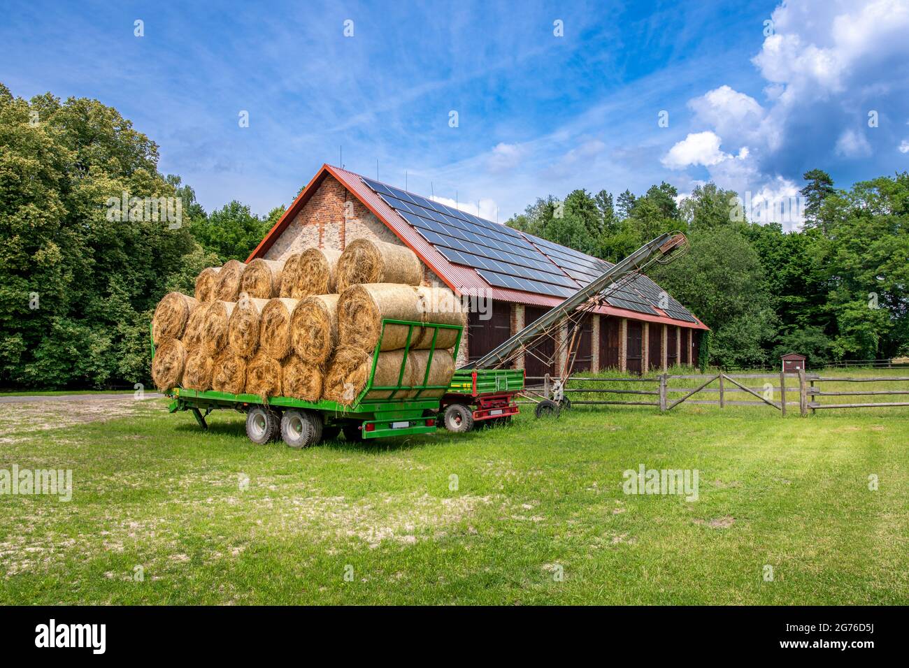Remorque verte remplie de balles de foin garées à l'avant de la grange en briques d'une ferme. Panneaux solaires installés sur le toit de la grange. Banque D'Images