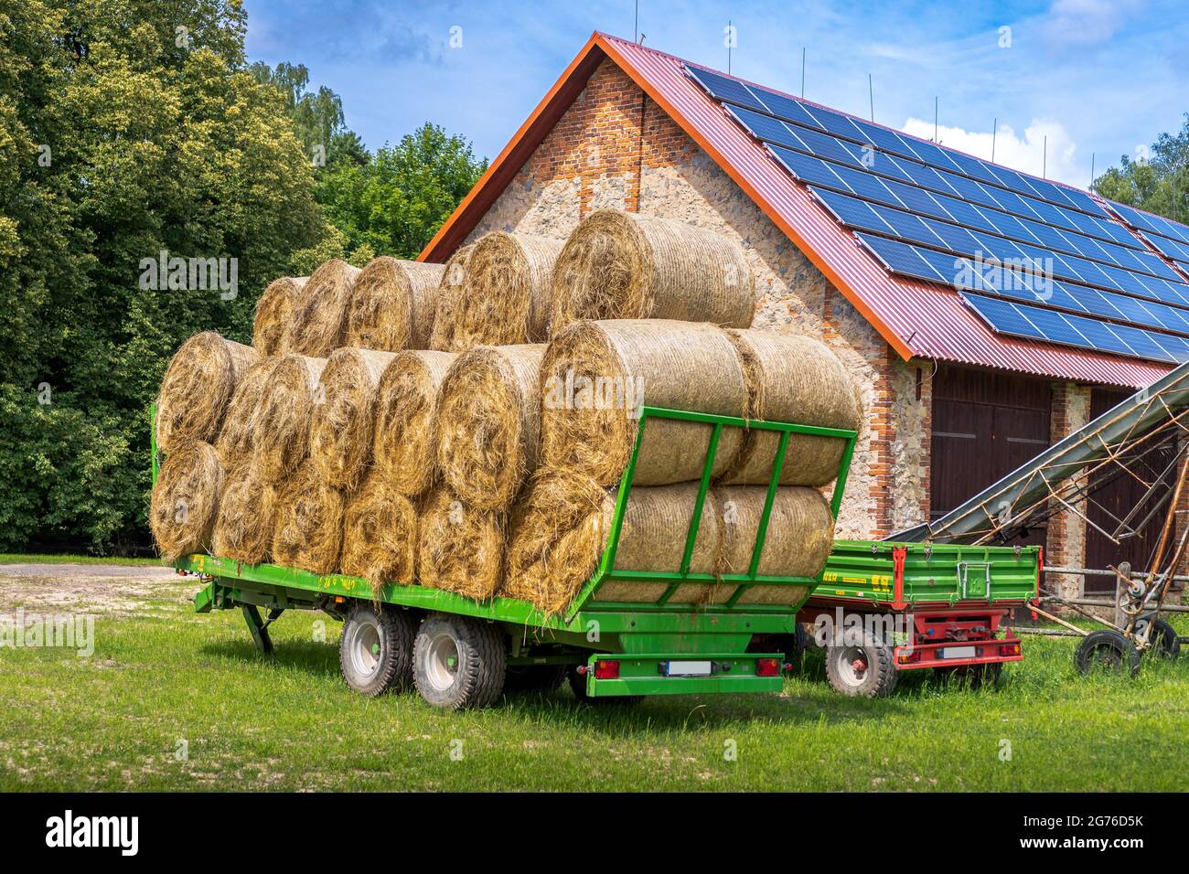 Remorque verte remplie de balles de foin garées à l'avant de la grange en briques d'une ferme. Panneaux solaires installés sur le toit de la grange. Banque D'Images