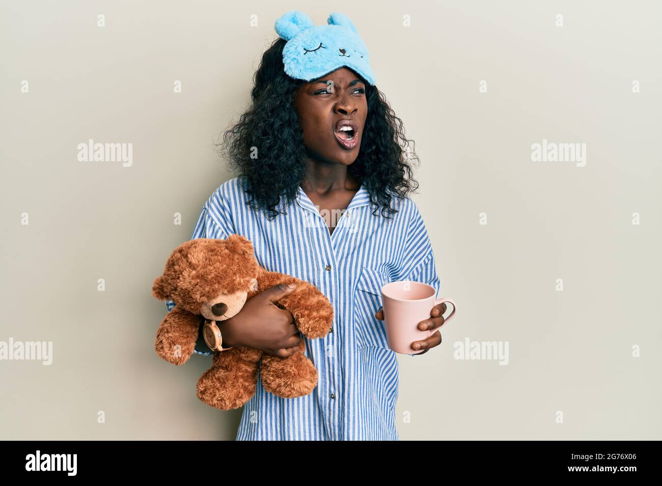 Belle jeune femme africaine portant un pyjama, tenant un ours en peluche et buvant un café en colère et fou hurlant frustré et furieux, criant avec ang Banque D'Images