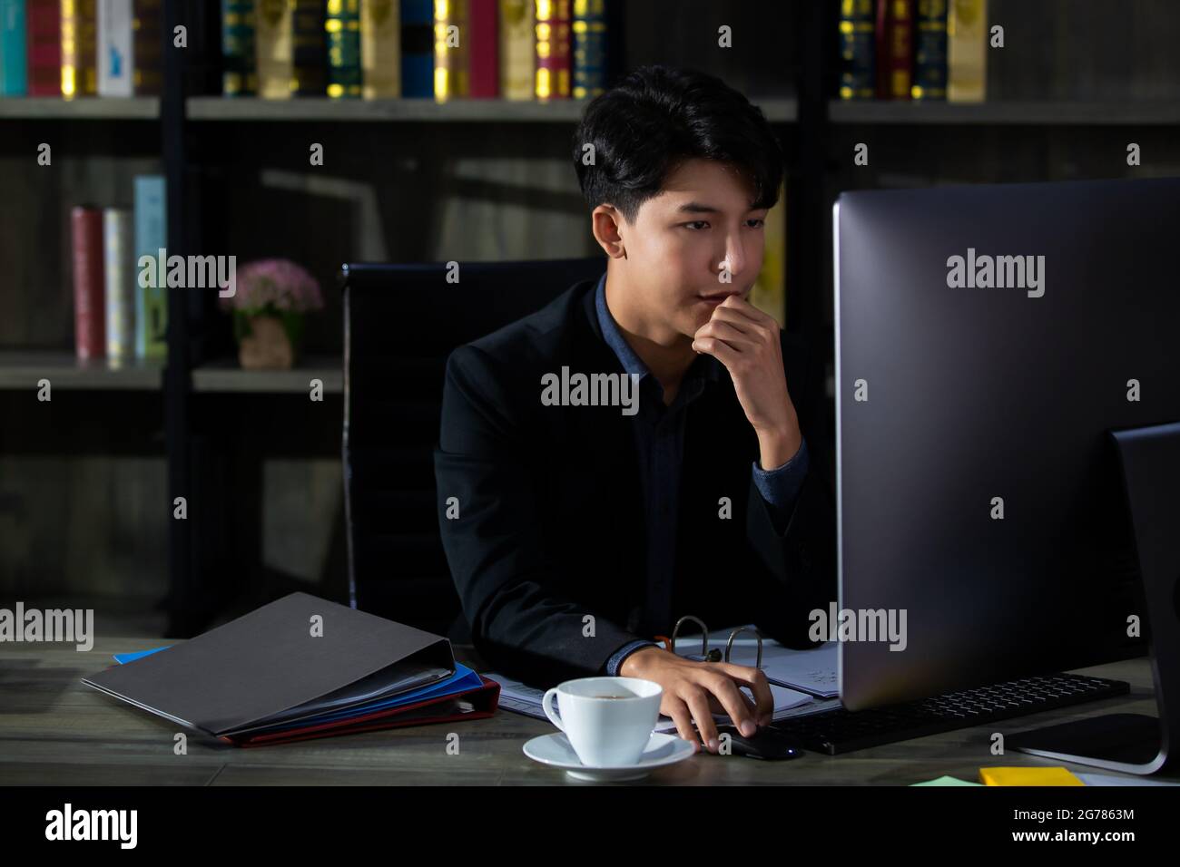Jeune et beau homme d'affaires asiatique nouvelle génération en costume assis dans la salle de travail privée et regardant l'écran d'ordinateur avec sérieux et réfléchi Banque D'Images