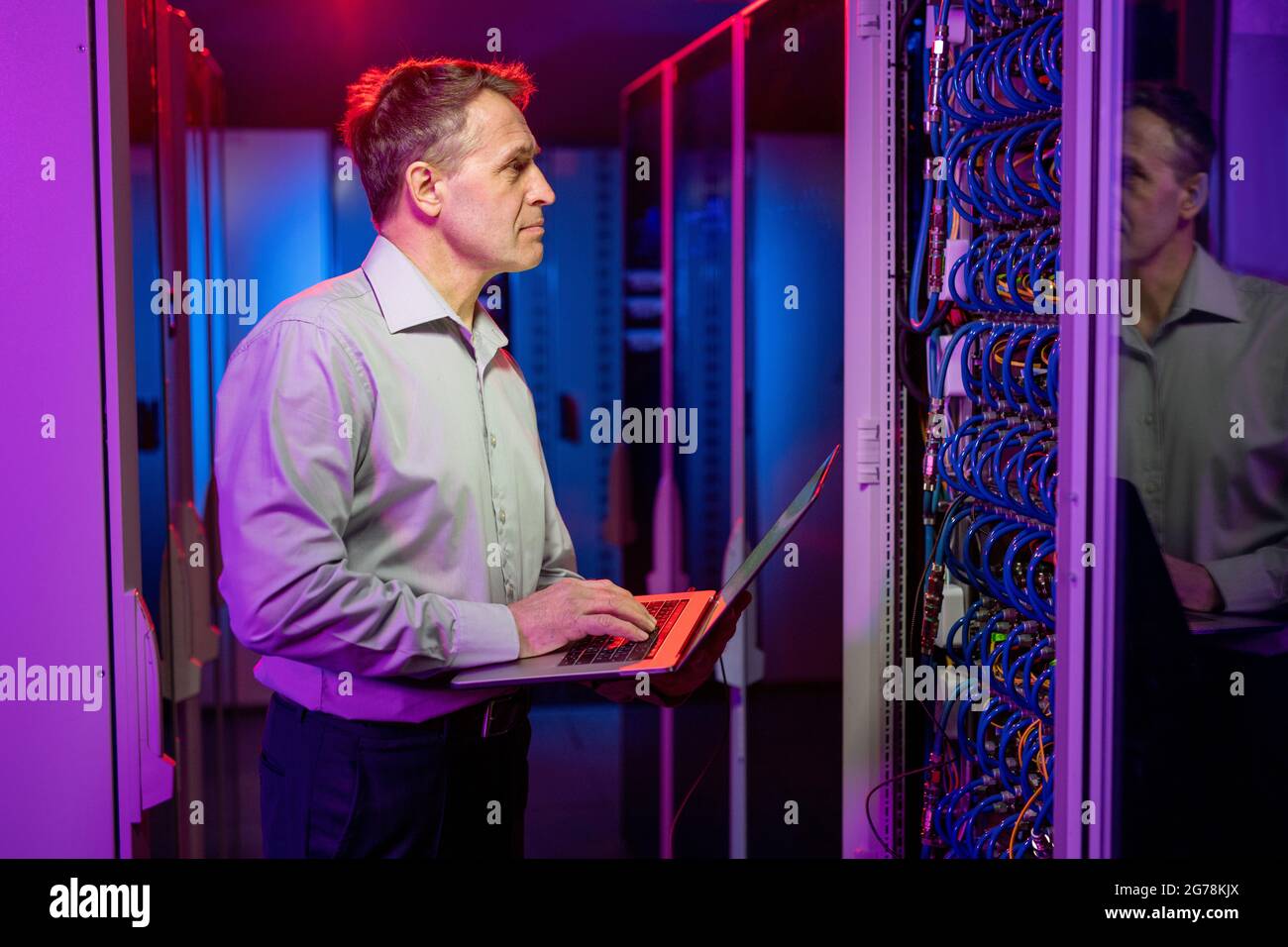 Ingénieur réseau caucasien mature concentré dans une chemise debout à l'armoire de serveur ouverte et effectuant un contrôle de sécurité à l'aide d'un ordinateur portable Banque D'Images