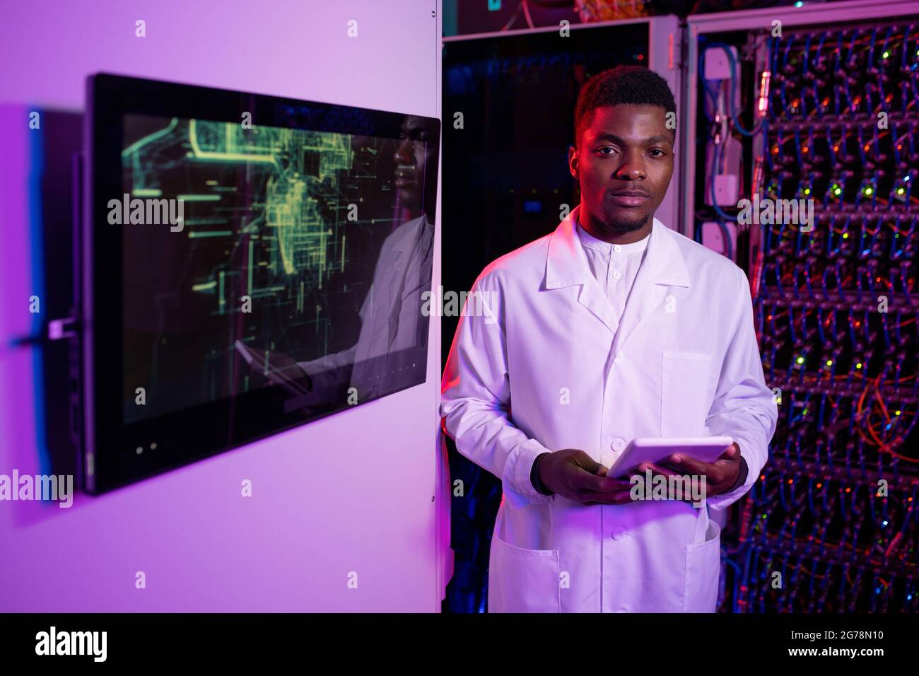 Portrait d'un jeune homme noir sérieux, en blanc, debout avec une tablette numérique contre une armoire de serveur dans la salle du centre de données Banque D'Images