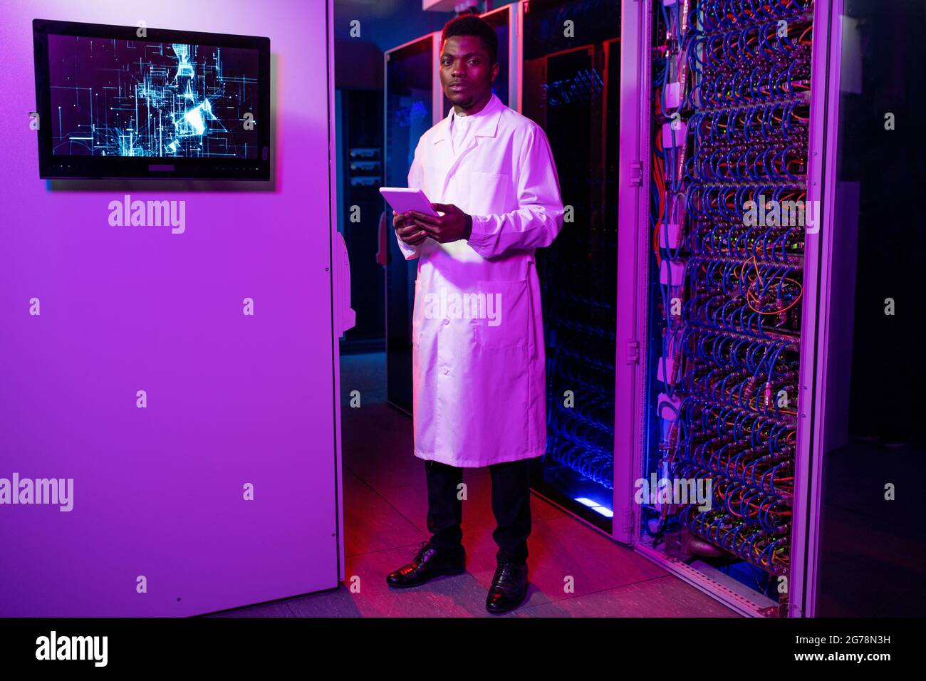 Portrait d'un jeune homme sérieux, afro-américain, administrateur INFORMATIQUE, en manteau blanc, debout avec une tablette dans une pièce au néon foncé avec des armoires Banque D'Images