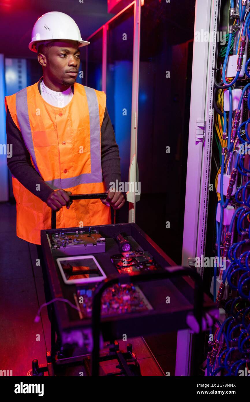 Jeune spécialiste sérieux de la réparation de serveur noir dans le chariot de poussée de casque et de gilet avec micropuces et cartes de circuit dans le centre de données en couleur néon Banque D'Images