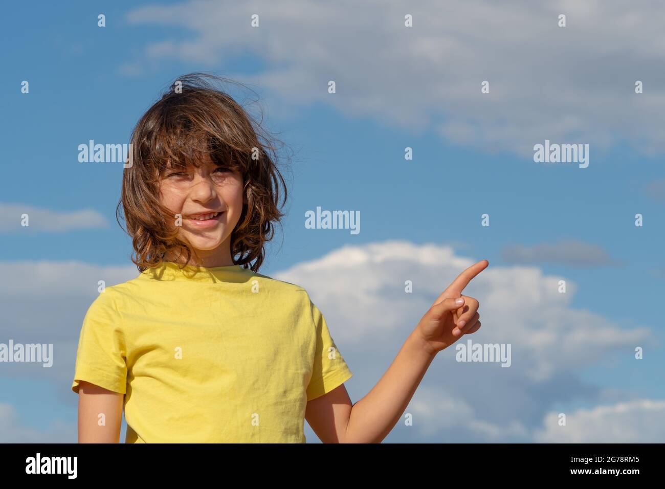 petit garçon souriant mignon avec de longs cheveux pointant vers le haut avec le doigt. ciel bleu avec fond de nuages. espace de copie Banque D'Images