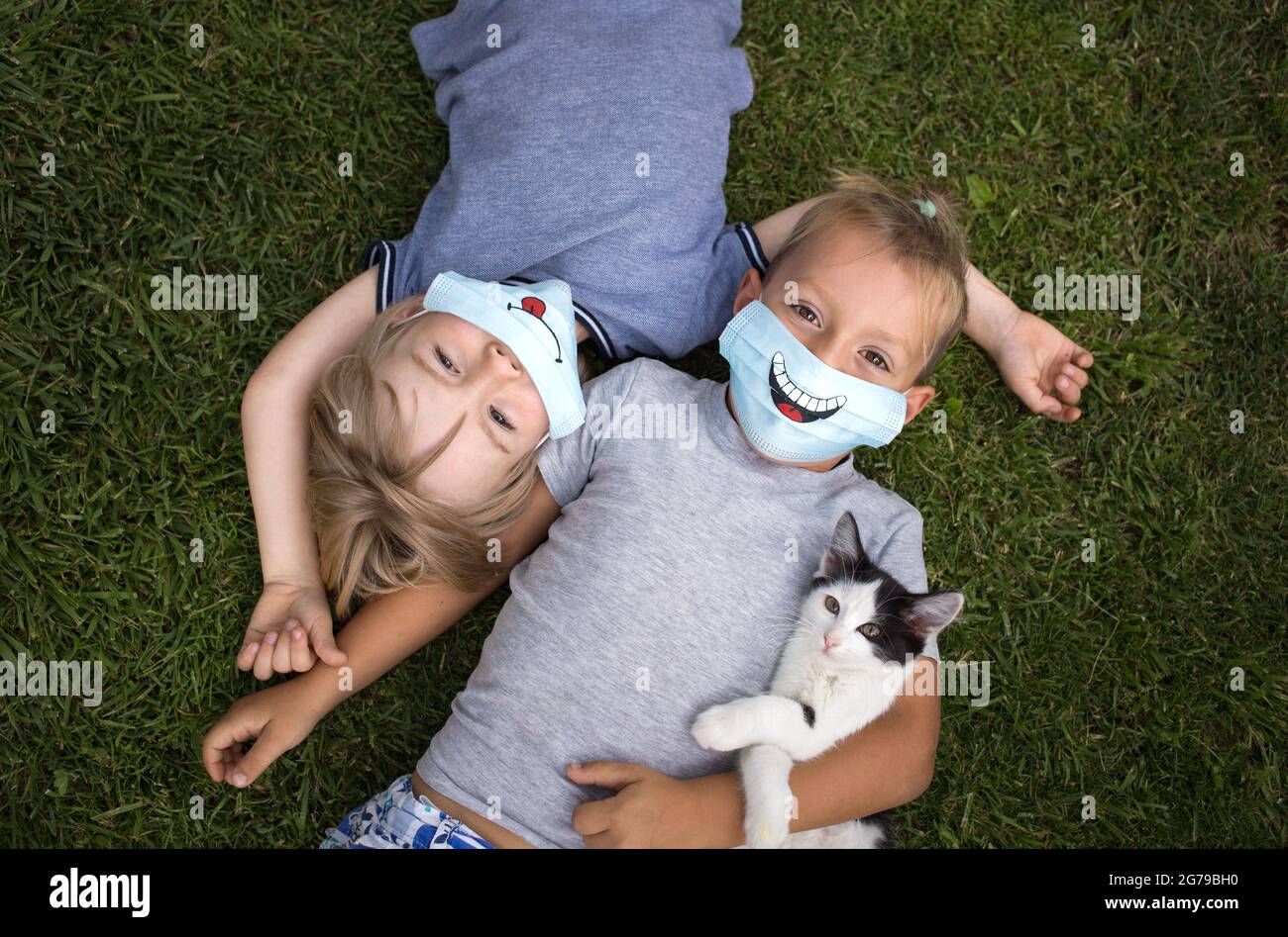 Deux tout-petits garçons se reposent sur le dos de l'herbe verte, regardant vers le haut. Les deux portent des masques de protection médicale bleus avec des sourires drôles. Communication entre les enfants Banque D'Images