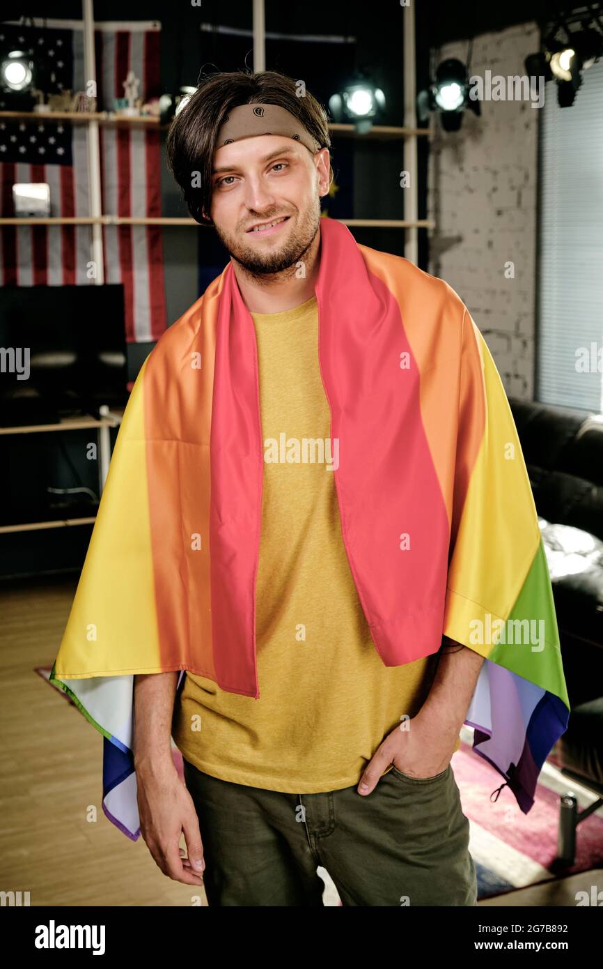 Un jeune homme heureux enveloppé dans un drapeau de fierté debout dans le salon de son appartement Banque D'Images