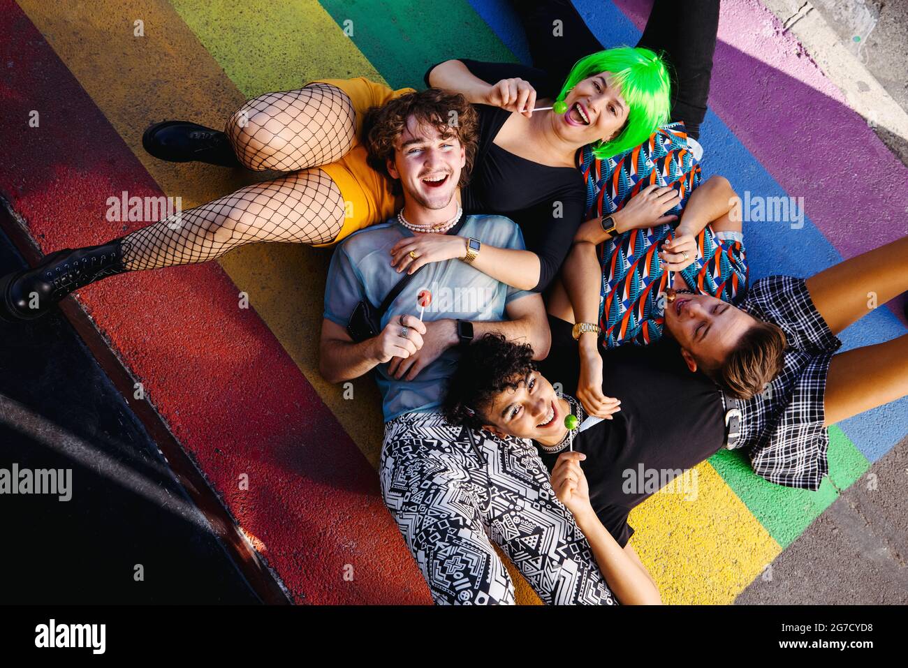 Les jeunes reines insouciants se couchés ensemble sur les couleurs de l'arc-en-ciel. Groupe de personnes heureux LGBTQ+ souriant gaiement pendant que chacun tient un lollipop. Ven Banque D'Images