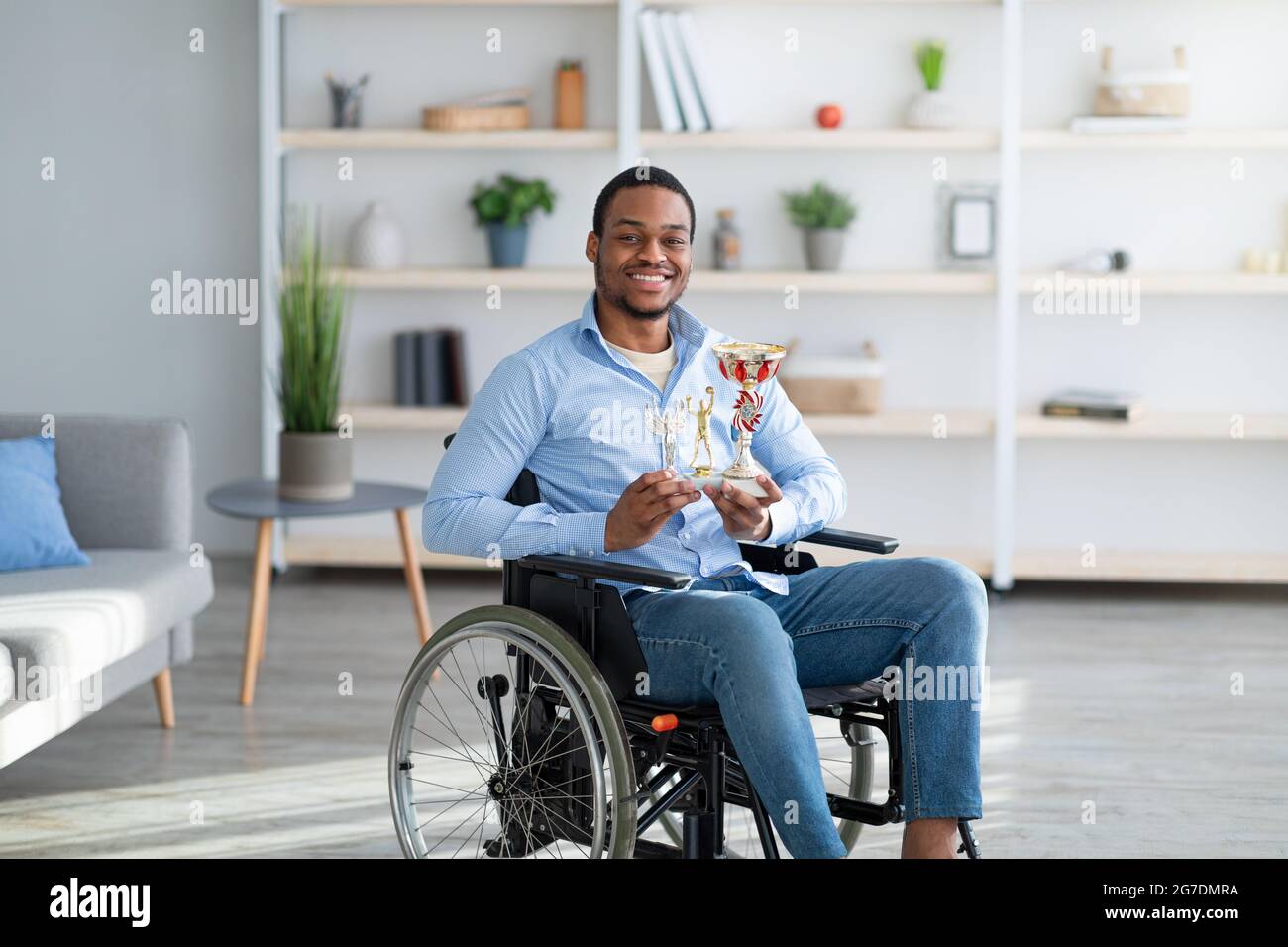 Joyeux sportif paraplégique en fauteuil roulant tenant des trophées et souriant à l'intérieur de la caméra Banque D'Images Joyeux sportif paraplégique en fauteuil roulant tenant des trophées et souriant à l'intérieur de la caméra Banque D'Images