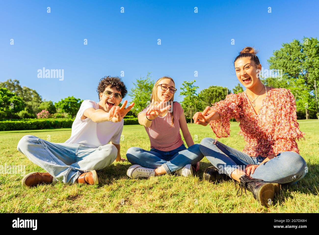 Trois jeunes étudiants meilleurs amis assis sur l'herbe dans le parc de la ville montrant le signe de victoire avec deux doigts regardant la caméra. Concept d'unité et de solidité Banque D'Images