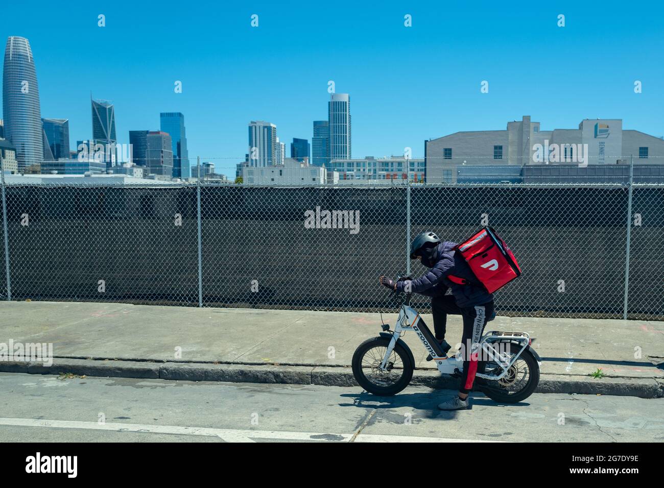 Conducteur de Doordash sur vélo électrique portant un sac à dos plus froid et téléphone de conseil avec horizon de San Francisco, Californie en arrière-plan, le 3 juin 2021. () Banque D'Images