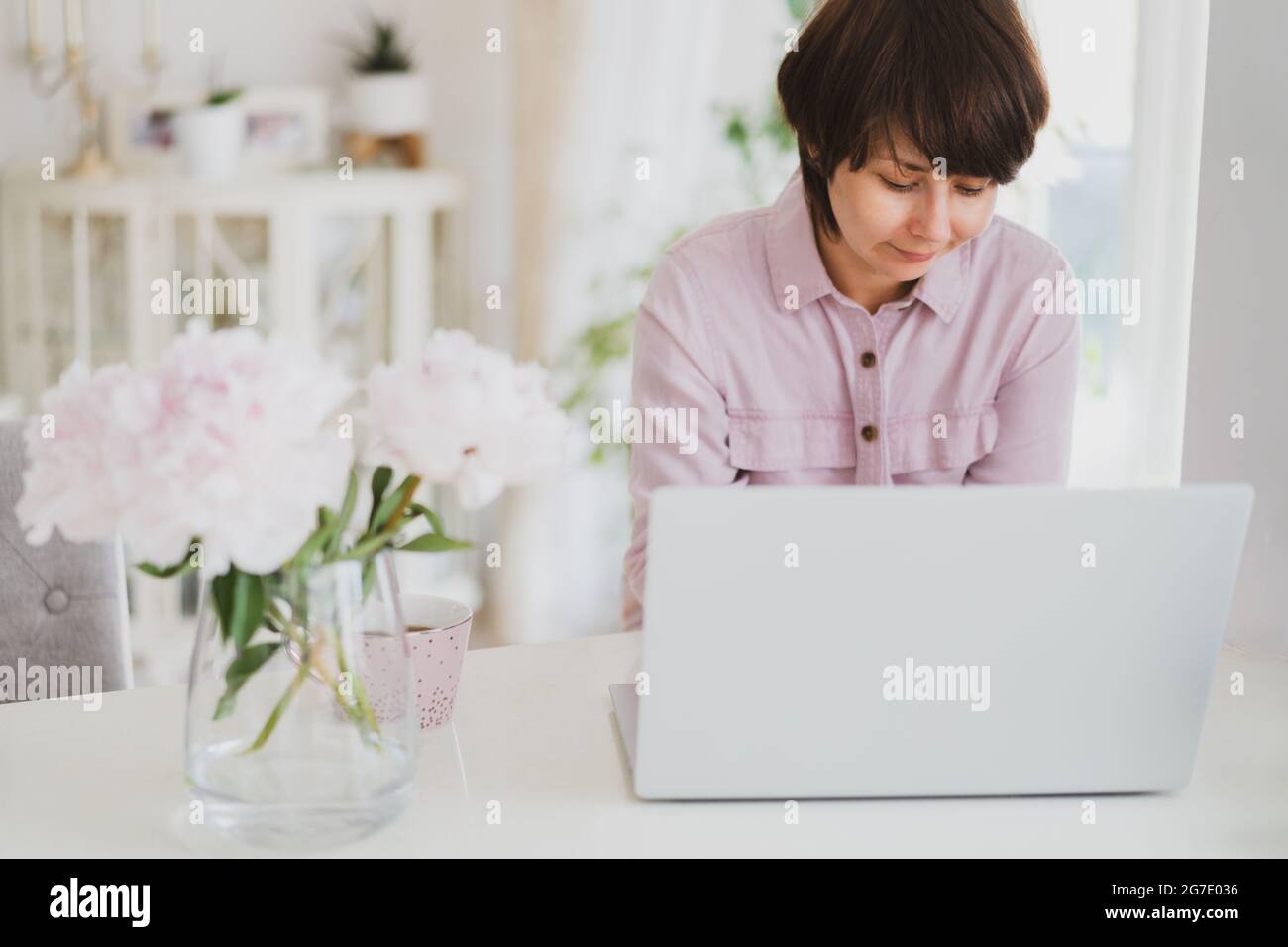 Femme de race mixte travaillant à la maison à l'aide d'un ordinateur portable. Espace de travail confortable avec fleurs de pivoines roses fraîches et une tasse de thé sur la table blanche avec lumière Banque D'Images