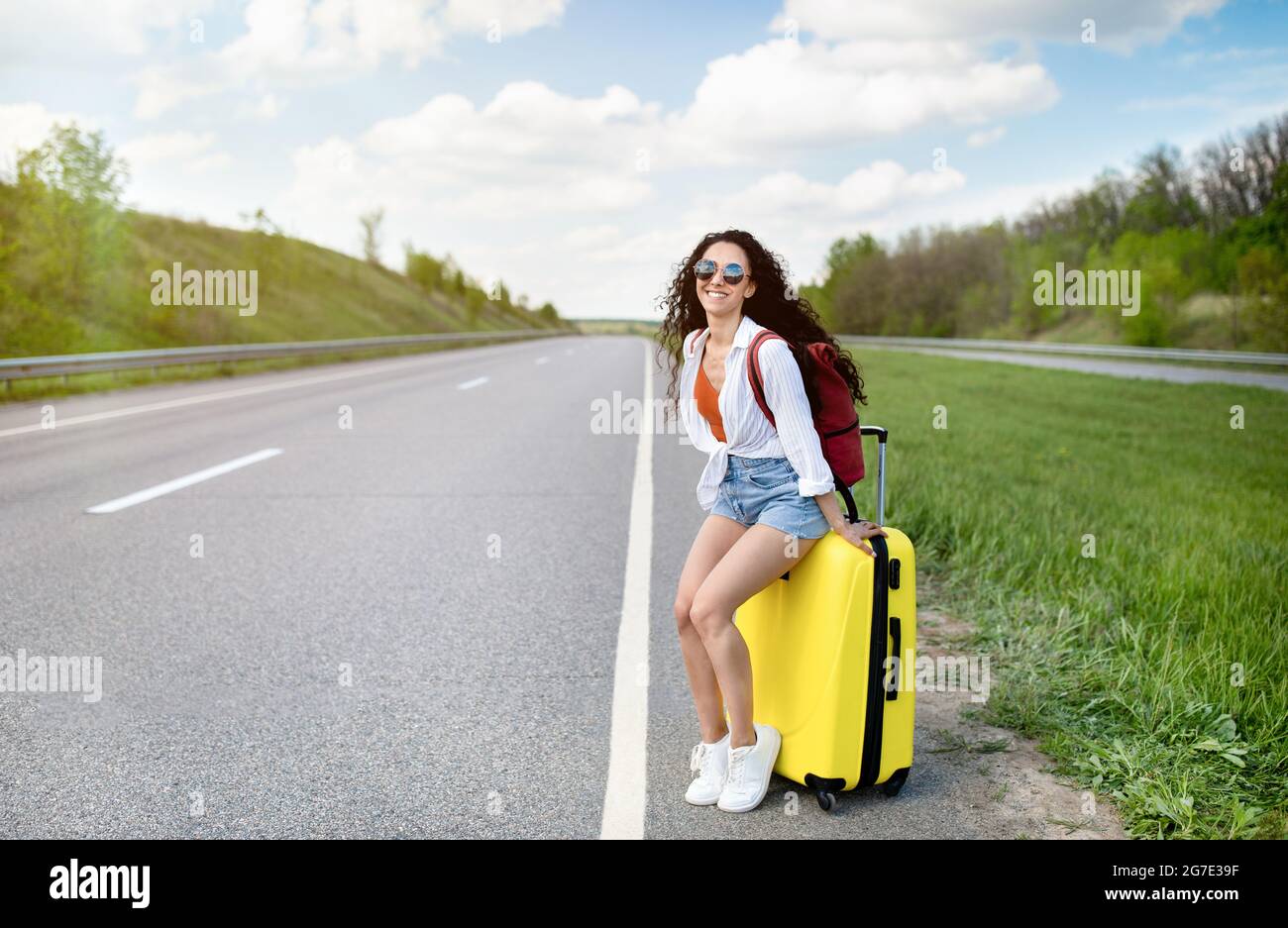 Jolie jeune femme assise sur une valise le long de la route, en faisant des promenades en voiture, en faisant des randonnées sur le bord de la route, en été Banque D'Images