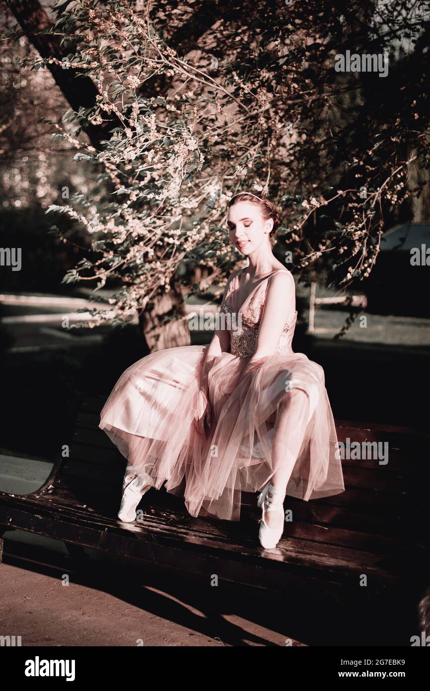 une ballerine dans une longue robe rose est assise à l'arrière d'un banc avec ses jambes larges à part avec ses pieds dans des chaussures de pointe sur la pointe en été Banque D'Images