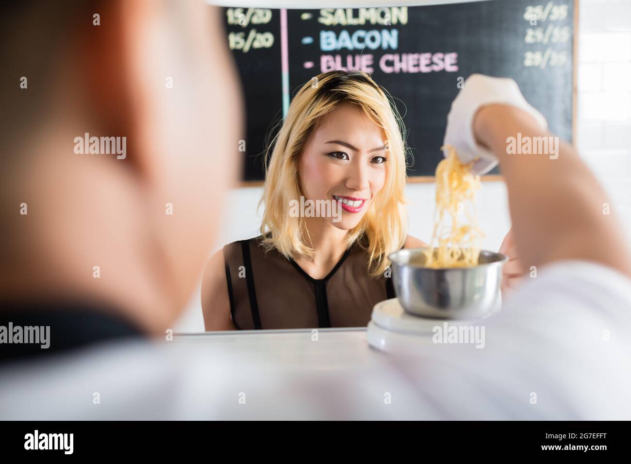 Femme asiatique regardant attentivement que le fromage râpé est placé sur une balance pour peser dans un restaurant ou deli dans une vue sur l'épaule de derrière le Banque D'Images