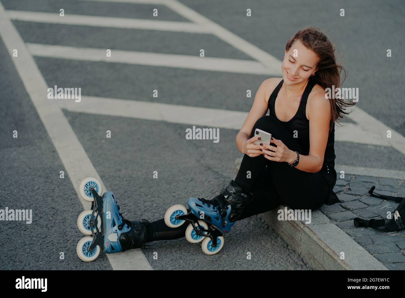 Vue de dessus de brunette active jeune femme utilise smartphone pour la communication en ligne poses sur l'asphalte vêtu de noir active port aime passe-temps favori Banque D'Images