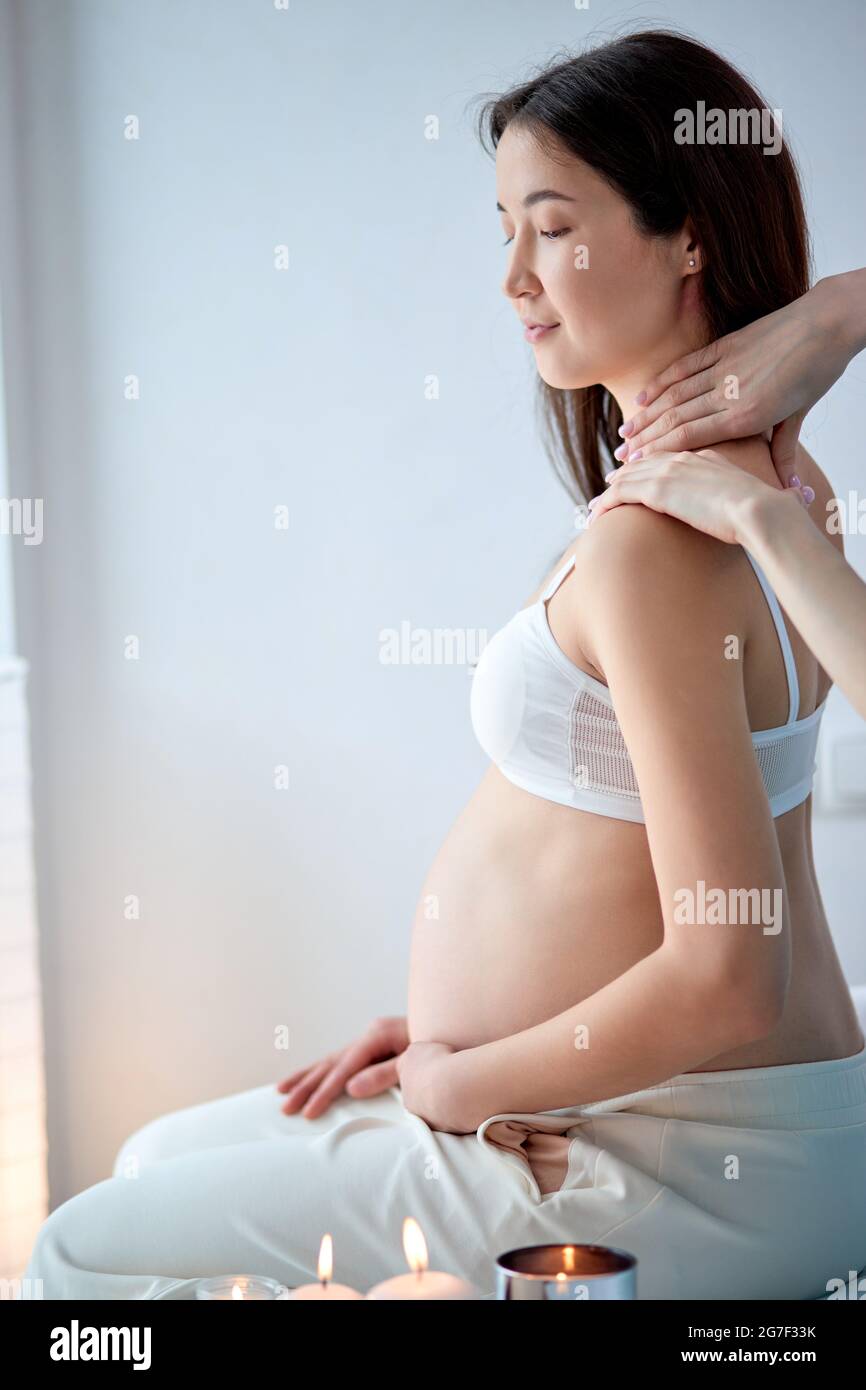 Jolie femme enceinte asiatique se massant au spa dans le dos, s'étreignant  le ventre, dans un soutien-gorge blanc. Vue latérale de la belle femme dans  le salon de spa en lumière Photo